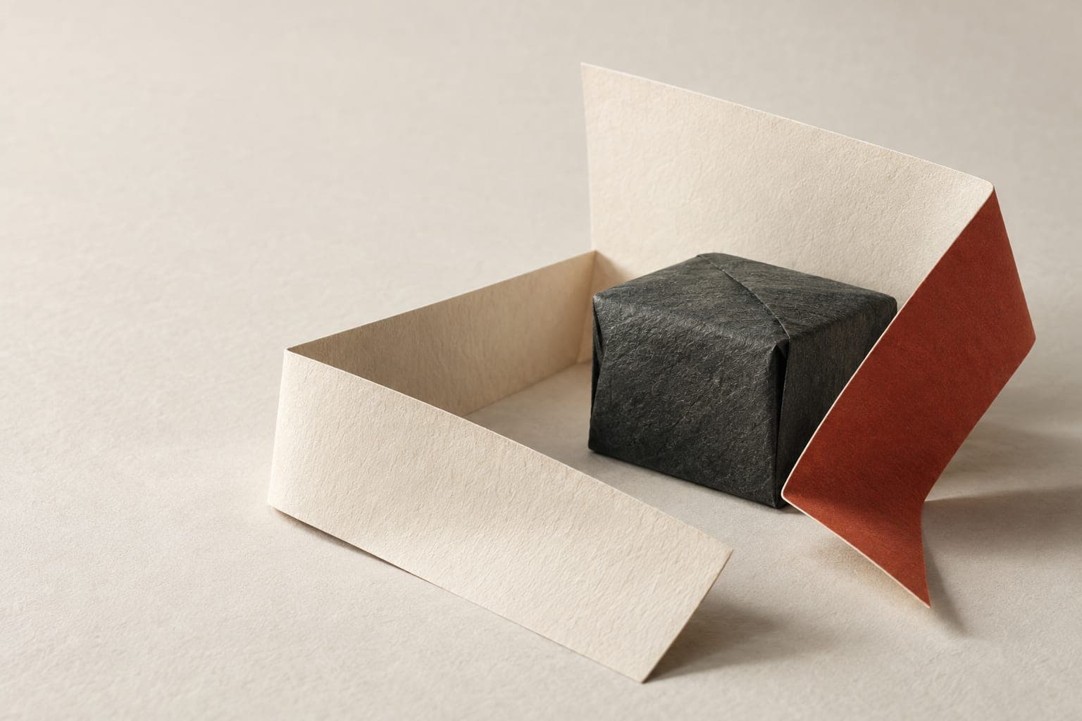 Folded cream paper surrounding a charcoal parcel-shaped object with a vermilion edge